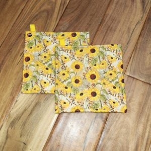 May include: Two square fabric pot holders with a yellow sunflower pattern. The pot holders have a yellow loop for hanging. The background is a wooden surface.