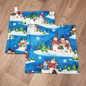 May include: Two blue quilted pot holders with a winter snowman theme. The pot holders feature snowmen, pine trees, and snowflakes on a blue background.