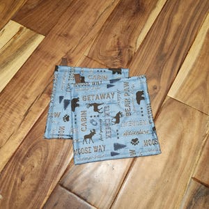 May include: Two blue and brown pot holders with a woodland theme. The pot holders feature a variety of designs, including moose, bear paw, elk creek, cabin, and moose way. The text on the pot holders reads "Cabin Mouse Way", "Wildlife Trail", "Getaway", "Nature Retreat", "Bear Paw", "Mountain Lodge", "Elk Creek", "Find Your Adventure", "Adventure", "Moose Way", "Wildlife Trail", and "Cabin".