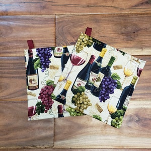 May include: Two white fabric pot holders with a wine theme. The pot holders feature images of wine bottles, grapes, and wine glasses. The pot holders have red fabric loops for hanging.