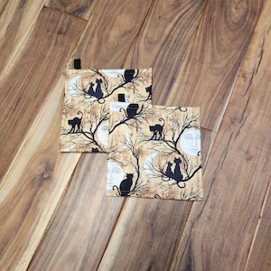 May include: Two black cat themed pot holders with a moon and tree design. The pot holders are made of a beige fabric with black cats, trees, and moons printed on them.