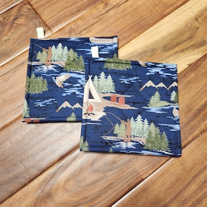 May include: Two blue pot holders with a camping and fishing theme. The pot holders feature a blue background with white, green, and brown accents. The design includes a tent, trees, a campfire, and a person fishing.