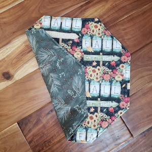 May include: A reversible fall-themed table runner. One side features a dark green fabric with a pattern of pine needles and berries. The other side has a black background with sunflowers, mason jars, and the word "FALL".