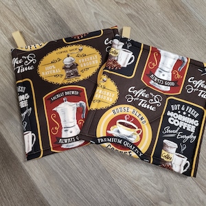 May include: Two square coffee-themed pot holders with brown, black, and yellow designs. The pot holders feature coffee-related text and images, including coffee cups, coffee beans, and coffee makers. Each pot holder has a tan loop for hanging.
