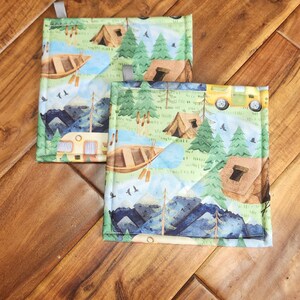 May include: Two square quilted pot holders with a camping theme. The fabric features a design of tents, trees, a river, a boat, and a camper. The pot holders have a green border and a small loop for hanging.