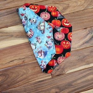 May include: Two fabric head coverings, one with a blue winter scene of snowmen and birds, the other with orange jack-o'-lanterns on a black background. The head coverings are folded and displayed on a wooden surface.