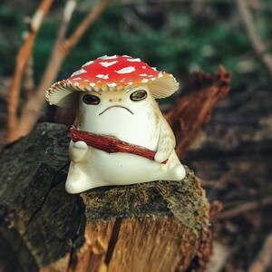 May include: A whimsical ceramic mushroom figurine with a red and white spotted cap and a grumpy expression. The mushroom character is holding a small, brown stick. The figurine is sitting on a weathered wooden surface.