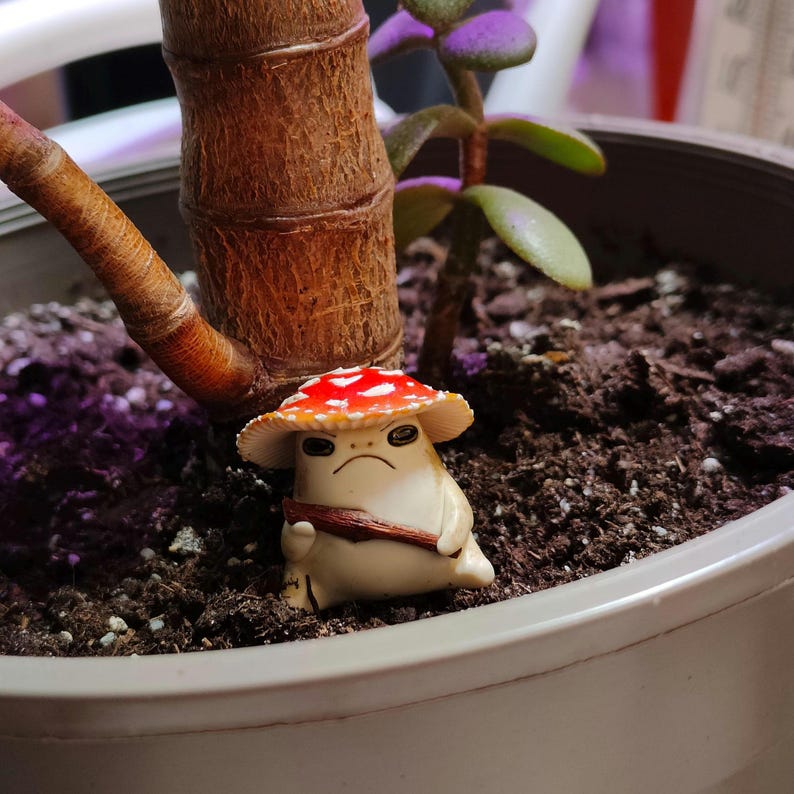 May include: A small, grumpy mushroom figurine with a red and white spotted cap and a brown sash. The figurine is set in a potted plant with a brown trunk and green leaves. The mushroom has a stern expression.