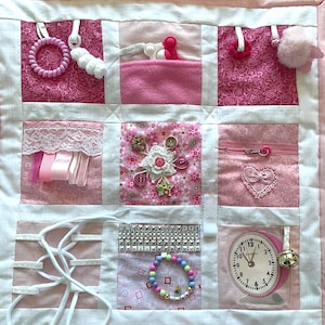 May include: A pink sensory activity quilt with various textures and interactive elements. Features include a zipper, buttons, ribbons, a bracelet, a clock, and a bell. The quilt is designed with a patchwork pattern and white borders.