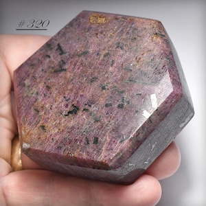 May include: A faceted, hexagonal gemstone with a deep pink and burgundy color, speckled with black inclusions. The stone is held in a hand, and the text "#320" is visible in the upper left corner.