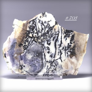 May include: A polished stone slab with a unique pattern of black, white, and pale purple hues. The stone has a natural, irregular shape and is mounted on a clear stand. The text "# 708" is visible on the upper right.