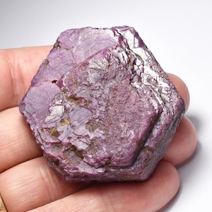 May include: A rough, faceted, deep magenta-colored mineral specimen. The surface has a crystalline texture with areas of light reflection. The stone is held in a hand, showcasing its size and shape. The overall appearance suggests a natural, unpolished gemstone.