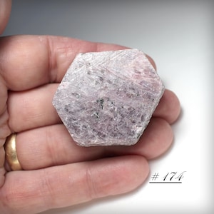 May include: A hexagonal, light pink gemstone with a rough, textured surface. The stone has dark gray inclusions and is held in a hand. The number "#174" is written in black script in the lower right corner.