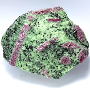 May include: A rough-cut stone featuring a vibrant green matrix speckled with black inclusions. Embedded within the stone are several elongated, deep pink ruby crystals. The combination of colors and textures creates a striking visual contrast.