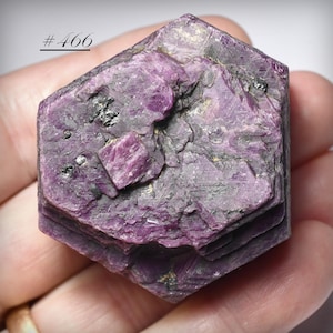 May include: A hexagonal, rough-hewn, deep magenta-colored mineral specimen. The surface has a layered appearance with dark gray and black inclusions. The text "# 466" is in the upper left corner. The mineral is held in a hand.