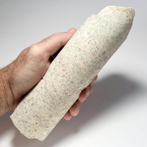 May include: A cylindrical geological core sample with a rough, speckled light gray and beige surface. The stone specimen features a dense, granular texture with visible mineral inclusions throughout its length, showing a natural, unpolished finish.
