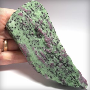 May include: A slab of zoisite mineral featuring a vibrant green base color with scattered inclusions of deep magenta ruby and dark black hornblende crystals. The stone has a rough, natural texture with an irregular, elongated triangular shape.