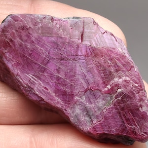 May include: A rough-cut, natural ruby gemstone with a deep magenta hue. The surface displays a network of fine, white veins and a crystalline structure. The stone has a raw, unpolished appearance, showcasing its natural beauty.