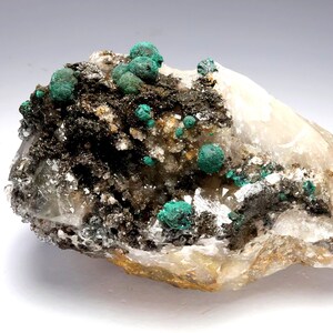 May include: A close-up of a mineral specimen featuring a light-colored base rock with dark, granular inclusions. Bright, spherical, teal-colored mineral formations are clustered across the surface, creating a striking contrast. The specimen has a rough, natural texture.