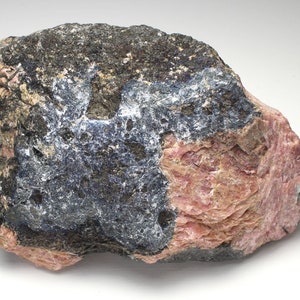 May include: A rough-hewn rock formation with a mix of colors. The rock features areas of dark gray, deep blue, and a prominent section of pinkish-red. The texture appears coarse and uneven, with visible mineral deposits.