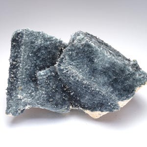 May include: A close-up of a mineral specimen featuring a cluster of blue-gray crystals. The crystals have a fibrous texture and are clustered on a light-colored base. The mineral has a rough, uneven surface.