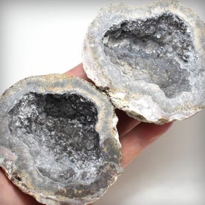 May include: Two halves of a geode, showcasing a hollow interior filled with sparkling crystals. The outer shell is a mix of gray and beige tones, with a rough, textured surface. The crystals inside glisten, creating a contrast with the muted exterior.