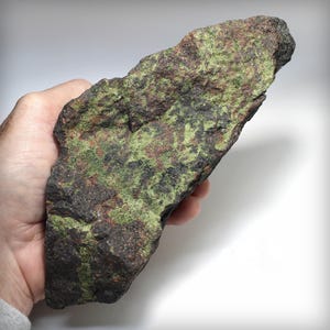 May include: A large, irregular rock specimen featuring a rough, textured surface with a mottled appearance. The stone displays a mix of deep reddish-brown, dark charcoal, and vibrant lime green mineral inclusions across its entire surface area.