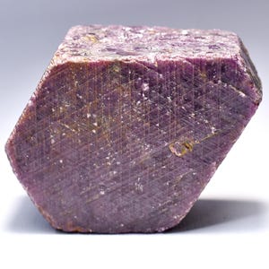 May include: A raw, hexagonal ruby crystal featuring a deep magenta and purple hue. The surface displays a distinct, natural geometric triangular pattern across its flat face, with visible crystalline inclusions and a rough, unpolished texture.