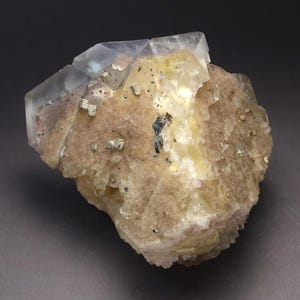 May include: A natural mineral specimen featuring a combination of clear, translucent crystals and a rough, beige matrix. The crystals have a geometric shape, and the matrix is speckled with small, metallic-looking inclusions. The overall color palette is a mix of clear, beige, and metallic.