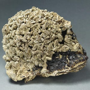 May include: A mineral specimen featuring a dense cluster of pale beige, tabular crystals covering a dark, metallic, cubic crystalline base. The structure displays a sharp contrast between the light-colored, fragmented surface and the dark, geometric mineral formation underneath.