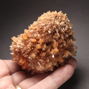 May include: A cluster of orange-brown crystals, resembling a geode or mineral formation. The crystals are pointed and clustered together, with a rough, textured surface. The overall shape is rounded and irregular, with a mix of light and dark tones.