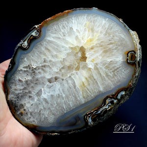 May include: A polished agate slice featuring a translucent, crystalline white center surrounded by concentric bands of dark brown, black, and grey. The outer edge is rough and textured, and the text PS1 is printed in white in the bottom right corner.