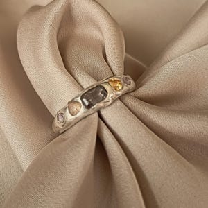 May include: A silver-toned ring with a textured, organic band featuring a central rectangular dark grey stone, flanked by two smaller teardrop-shaped stones in yellow and light brown, and two tiny round light purple stones on the outer edges.