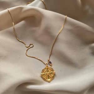 Op de afbeelding: Een goudkleurige ketting met een kruisvormige hanger. De ketting heeft een fijne ketting en een kleine paarse edelsteenbedel. De hanger is gedetailleerd met ingewikkelde patronen en texturen, tegen een zachte, crèmekleurige achtergrond.