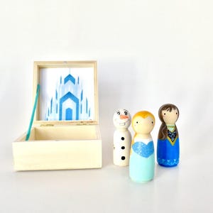 K&ouml;nnte beinhalten: Eine Holzkiste mit einer blau-wei&szlig;en Schlossillustration im Inneren, zusammen mit drei bemalten Holzpuppen: einem Schneemann, einer blonden Puppe in einem blauen Kleid und einer br&uuml;netten Puppe in einem blauen Kleid.