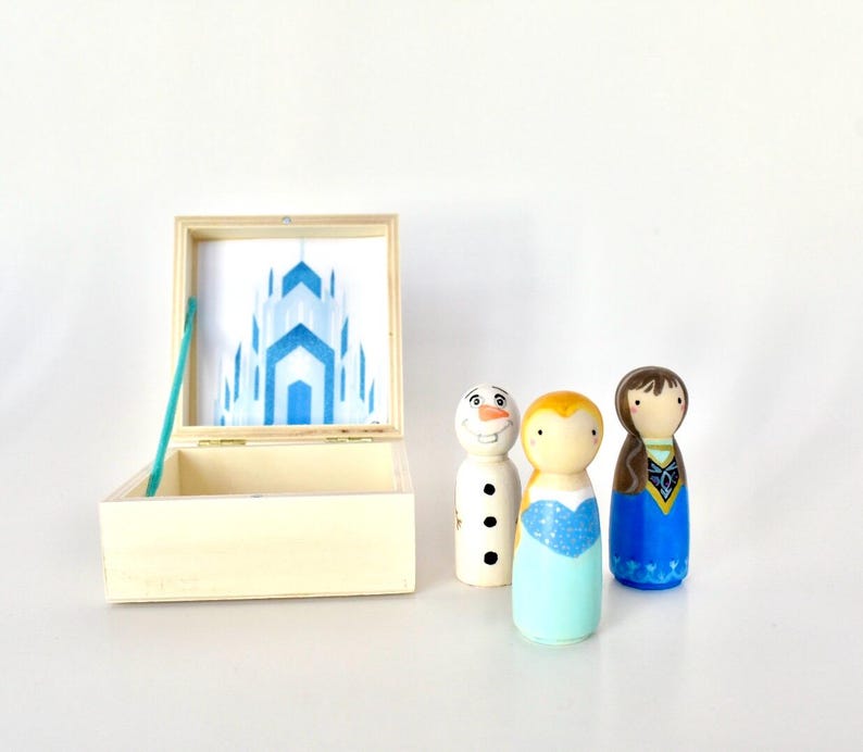 K&ouml;nnte beinhalten: Eine Holzkiste mit einer blau-wei&szlig;en Schlossillustration im Inneren, zusammen mit drei bemalten Holzpuppen: einem Schneemann, einer blonden Puppe in einem blauen Kleid und einer br&uuml;netten Puppe in einem blauen Kleid.