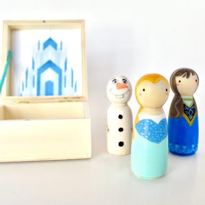 K&ouml;nnte beinhalten: Handbemalte Holzfiguren von Charakteren aus dem Film Frozen, darunter Olaf, Anna und Elsa. Die Figuren stehen neben einer Holzkiste mit einem blauen Schlossdesign. Die Kiste ist ge&ouml;ffnet.