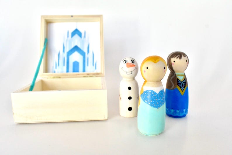 K&ouml;nnte beinhalten: Handbemalte Holzfiguren von Charakteren aus dem Film Frozen, darunter Olaf, Anna und Elsa. Die Figuren stehen neben einer Holzkiste mit einem blauen Schlossdesign. Die Kiste ist ge&ouml;ffnet.