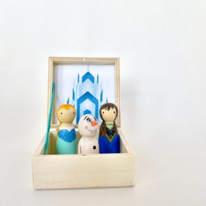 K&ouml;nnte beinhalten: Holzkiste mit einem bemalten Hintergrund eines blauen Schlosses und drei Holzpuppen. Die Puppen sind bemalt, um Figuren aus einem beliebten Animationsfilm darzustellen. Die Kiste ist ge&ouml;ffnet und zeigt die Puppen und die Schlossszene.
