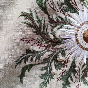 Puede incluir: Primer plano de un diseño bordado blanco y verde sobre una tela beige. El diseño presenta una gran flor blanca con un centro marrón y hojas verdes.