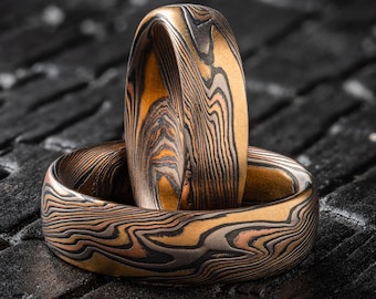 May include: Two handcrafted rings with a unique, layered pattern in shades of gold, copper, and black. The rings are set against a dark, textured background, highlighting their intricate design and metallic sheen. The rings appear to be made of metal.