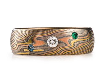 May include: A gold band ring with a patterned surface and three gemstones. The ring has a large, round, white gemstone in the center, a smaller, round, blue gemstone on the left, and a smaller, round, green gemstone on the right.