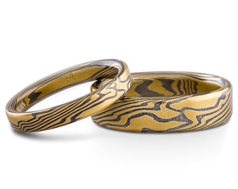 May include: Two gold wedding bands with a black and gold patterned design.