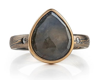 Puede incluir: Un anillo en forma de pera con una piedra preciosa gris oscura engastada en un bisel de oro. La banda está hecha de metal negro y plateado con un acabado texturizado y presenta dos pequeños diamantes.