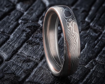 May include: A Damascus steel ring with a distinctive pattern. The ring displays a mix of silver, copper, and dark grey tones, creating a layered, wood-grain effect. The ring is set against a dark, textured background.