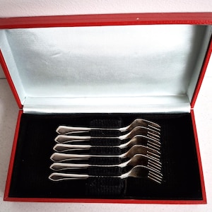 Puede incluir: Un juego de seis tenedores plateados con mangos decorativos, presentados en una caja roja con interior de terciopelo negro y forro de satén blanco. Los tenedores están dispuestos ordenadamente, mostrando su elegante diseño.