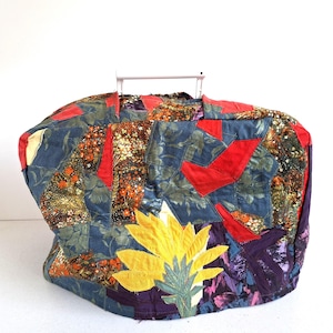 Puede incluir: Un bolso tote de patchwork con asa blanca. La bolsa presenta una variedad de patrones de tela, incluyendo estampados florales y colores lisos. Los colores dominantes son el azul, el rojo, el amarillo y el morado. Una gran flor amarilla está aplicada en la parte delantera.
