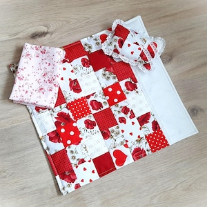 Peut inclure: Une couverture patchwork faite à la main pour bébé avec un oreiller assorti et un tissu plié. La couverture présente des carrés rouges et blancs avec des motifs floraux, à pois et en forme de cœur. L'oreiller a une bordure en dentelle et des détails en forme de cœur.