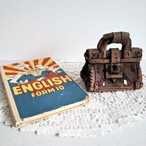 Könnte beinhalten: Ein altes Lehrbuch English Form 10 mit blau-gelbem Einband und Comic-Illustration, neben einer kleinen, braunen, texturierten Tonskulptur in Form einer strukturierten Handtasche mit einem zentralen Schnallendetail.