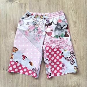 Op de afbeelding: Patchwork shorts met verschillende patronen, waaronder roze stippen, bloemenprints en vlindermotieven. Een grijze koala beer staat op één been. De shorts hebben een elastische tailleband en zijn gemaakt van zachte stof.